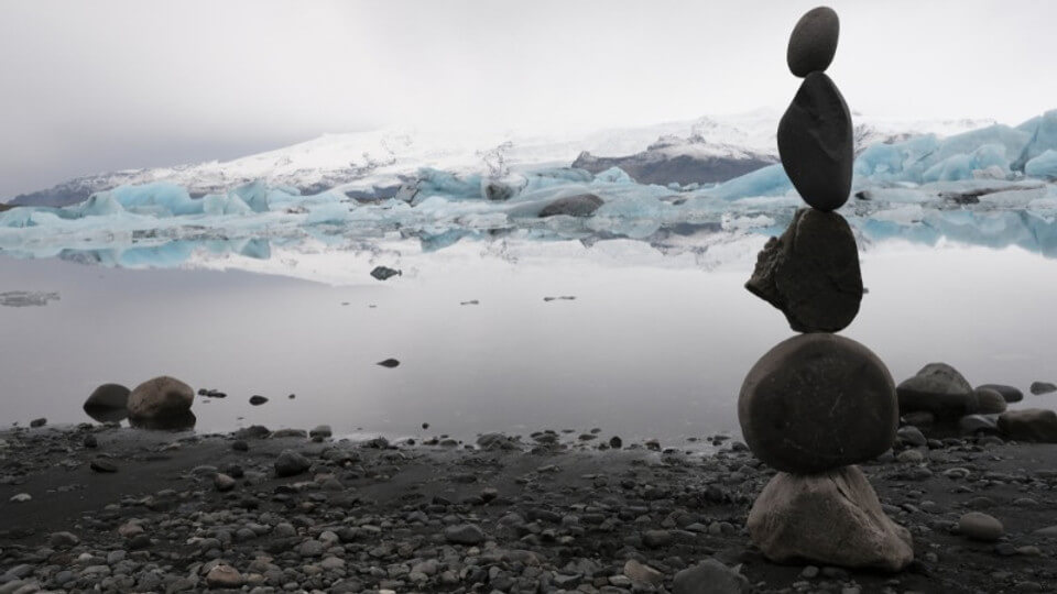 Stacked rocks balancing on the shore of a serene glacial lake with icebergs and snow-covered mountains reflecting in the still water in Iceland.
