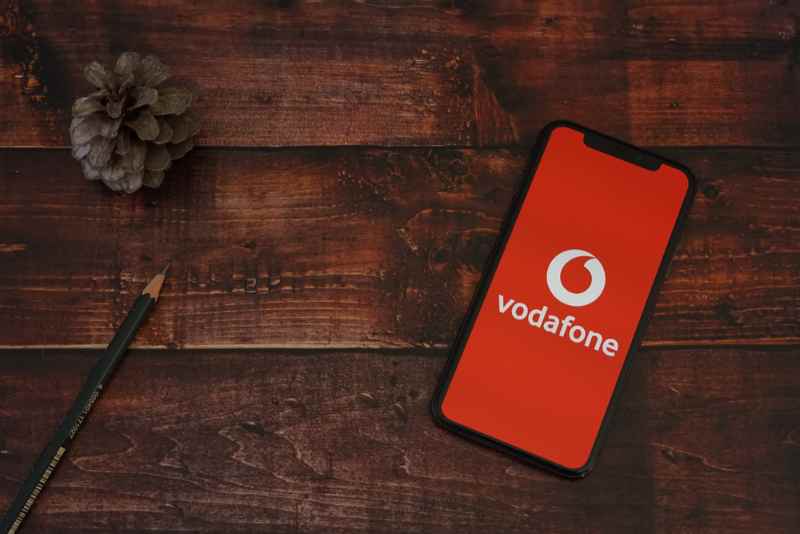 Vodafone logo on a smartphone lying on a rustic wooden table Smartphone with the Vodafone logo on the screen lying on a wooden table next to a pencil and a pine cone.