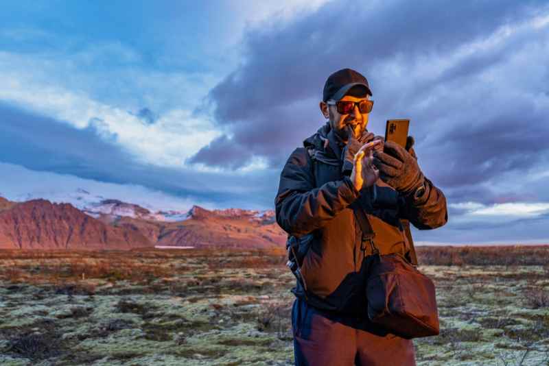 Man with smartphone in front of a breathtaking Icelandic landscape at sunset Man in outdoor clothing using his smartphone in front of a picturesque landscape in Iceland during sunset.