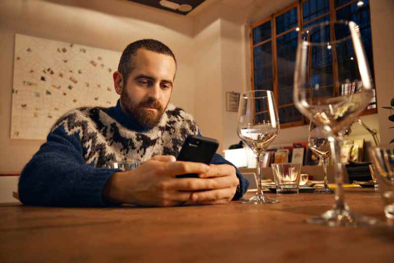 Man in Icelandic sweater uses smartphone in cozy restaurant Man in traditional Icelandic sweater sitting at a table in a restaurant and using his smartphone, surrounded by wine glasses.