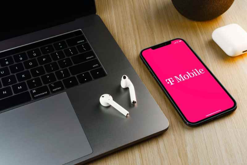 T-Mobile smartphone next to laptop and wireless headphones Smartphone with T-Mobile logo on the screen next to a laptop and wireless headphones on a wooden table.